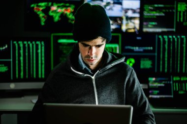 Genç hacker, yeşil kod gösteren birden fazla bilgisayar ekranıyla çevrili ev ofisinden kurumsal sunuculara girmeye odaklanıyor.