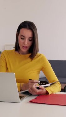 Evde dizüstü bilgisayar kullanırken not alan genç bir kadının dikey HD videosu. Milenyum güzeli kadın çevrimiçi sanal video dinlemeyi öğreniyor. İş ve eğitim kavramı