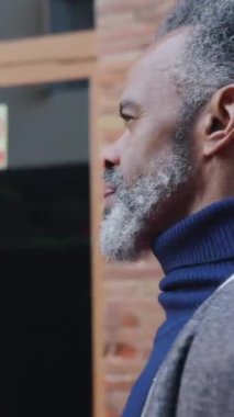 Dikey HD video: şehir caddesinde modern ofis binasının dışında yürüyen kıdemli Afrikalı Amerikalı iş adamının yan görüntüsü. O 60 'lı yaşlarda. Olumlu duygular, yaşlı insanlar.