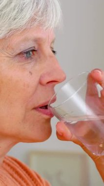 Dikey HD video. Sabah uyandıktan sonra evde kadehten su içen yaşlı bir kadın var. Sağlıklı yaşam tarzı kavramı.