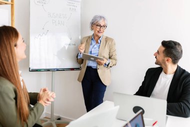 Modern bir ofiste ekibiyle birlikte bir iş sunumunu yönetirken dijital tablet kullanan kıdemli iş kadını. İş ve takım çalışması kavramı