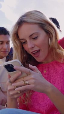 Sosyal medya içeriğini cep telefonu uygulamasından izleyen genç bir arkadaş grubunun dikey HD videosu. Milenyum çeşitliliği olan insanlar dışarıda otururken akıllı telefon kullanıyor. Gençlik ve teknoloji kavramı