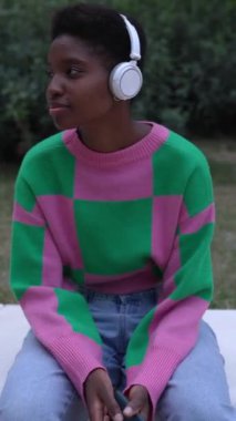 Halk parkında otururken binlerce yıllık neşeli Afrikalı kadının çevrimiçi müzikten zevk aldığı dikey HD video. Siyah kadın radyo yayını dinliyor ya da açık havada dinlenirken en sevdiği şarkı