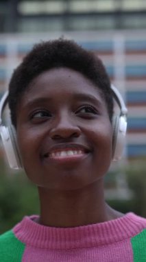 Kulaklıklı, şehir sokaklarında dikey duran genç siyahi kadının dikey HD videosu..