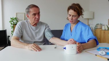 Young female healthcare worker measuring arterial blood pressure of senior man sitting on living room table at home. Hypertension in older age and medical home assistance concept.