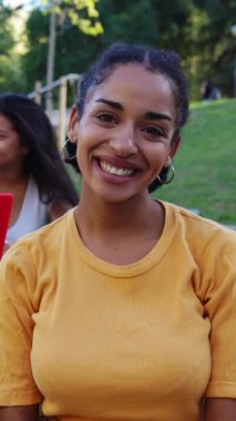 Genç Latin Gen Z öğrencisinin dikey portresi, kameraya gülümseyen kız ve üniversite kampüsünde dizüstü bilgisayarda çalışan sınıf arkadaşlarıyla birlikte oturuyor. Eğitim ve üniversite yaşam tarzı konsepti