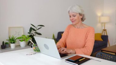 Odaklanmış yaşlı kadın evde çalışıyor, masada oturuyor ve dizüstü bilgisayarında yazıyor. İş ve telekomünikasyon kavramı