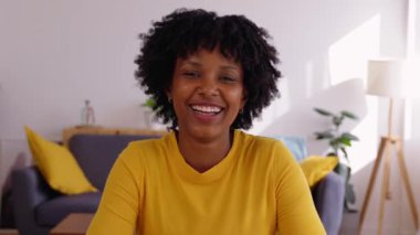 Oturma odasındaki masada oturan, kameraya gülümseyen neşeli, genç siyahi bir kadının portresi. Web kamerası görünümü veya POV