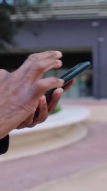 Dikey video. Yaşlı Afro-Amerikan bir adam şehir caddesinde yürürken cep telefonu kullanıyor. Olgun emekli erkek akıllı telefon uygulamasında çevrimiçi mesaj gönderiyor veya sosyal medya içeriğini araştırıyor. 