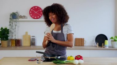 Neşeli, genç, çok kültürlü bir kadın modern mutfakta taze sebzelerle sağlıklı bir yemek hazırlarken tahta kaşığı mikrofon olarak kullanarak şarkı söylerken eğleniyor. Boş zaman ve ev hayatı konsepti