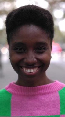 Şehrin arka planında kameraya gülümseyen genç ve mutlu bir Afrikalı kadının dikey HD videosu. Açık havada duran siyah bir kadının vesikalığı..