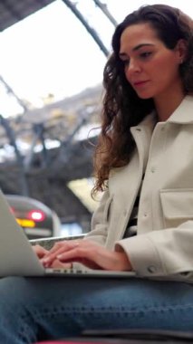 İstasyonda treni beklerken dizüstü bilgisayarda çalışan genç bir kadının dikey HD videosu. Seyahat ve iş insanları yaşam tarzı konsepti