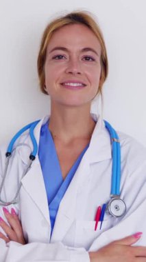 Beyaz arka planda kollarını kavuşturmuş kendine güvenen genç bir doktorun gülümseyen portresi. Mavi üniforma ve beyaz laboratuvar önlüğü giyiyor. Sağlık hizmetleri insanları konsepti. Dikey video.