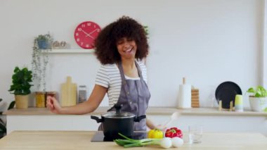 Modern mutfakta taze sebzelerle sağlıklı çorba pişirirken şarkı söyleyip dans eden neşeli, çok kültürlü genç bir kadın. Boş zaman ve ev hayatı konsepti