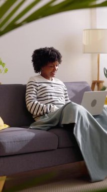 Mutlu, güzel, mutlu, Afro-Amerikan kadın kış günü evinde kanepede dinlenirken rahat bir battaniyeye sarılıp dizüstü bilgisayar kullanıyor. Teknoloji ve iş konsepti. Dikey HD video.