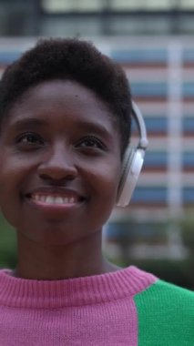 Kulaklıklı, şehir sokaklarında dikey duran genç siyahi kadının dikey HD videosu..