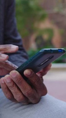 Dikey video. Yaşlı bir zenci, şehir caddesinde otururken cep telefonu kullanıyor. Tanımlanamayan yetişkin erkek çevrimiçi mesaj gönderiyor ya da sosyal medya içeriği içerikli cep telefonu uygulaması arıyor. 