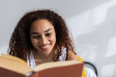 Genç mutlu Latin Amerikalı kadın yatak odasında kitap okuyor, doğal ışıkla aydınlatılıyor.