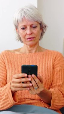 Dikey HD video. Evdeki koltukta oturmuş akıllı telefon mesajları okuyan endişeli yaşlı kadın..