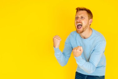 Heyecanlı adam zaferi kutlarken yumruklarını kaldırmış ve ağzı açık, canlı sarı bir arka plana neşe saçıyor.