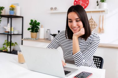 Genç ve mutlu bir kadın sabah kahve içerken mutfaktaki dizüstü bilgisayarda çalışıyor. İnternette öğrenen kız öğrenci. Eğitim ya da iş konsepti