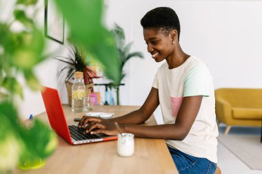 Genç, mutlu Afrikalı bir kadın bilgisayarla evde yoğurt yerken internetten öğreniyor.