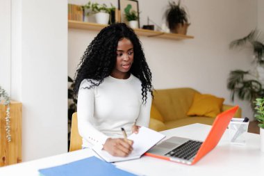 Genç siyahi kadın dizüstü bilgisayarını kullanarak uzaktan çalışırken not defterine not alıyor. Girişimciler ve iş konsepti