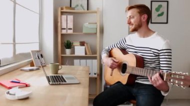 Mutlu genç adam evden bilgisayarla uzaktaki öğretmeniyle online müzik dersinde gitar öğreniyor. Genç yetişkinler dizüstü bilgisayarla müzik öğretiyor. Uzaklık eğitimi öğrenme kavramı