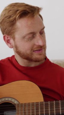 Neşeli genç kızıl adam yeni evindeki kanepede gitar çalıyor. Müzik, taşınma ve taşınma konsepti. Dikey HD portre videosunu kapat