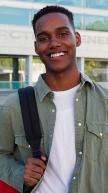 Genç Afro-Amerikan Gen Z öğrencisinin üniversite kampüsünün önünde dikey HD video portresi. Sırt çantalı neşeli genç dışarıda kameraya gülümsüyor.