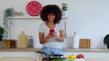 Mutfakta yemek pişirirken cep telefonu kullanan, tavada biraz sebze kızarmasını bekleyen mutlu, çok ırklı genç bir kadın. Teknoloji ve sağlıklı yaşam tarzı kavramı