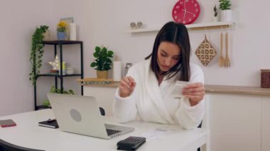 Bornozlu genç bir kadın elinde kağıt para ile mutfaktaki masada oturuyor, ev ekonomisini yönetiyor ve aylık faturaları hesaplıyor.