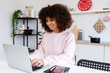 Genç mutlu Afro-Amerikan kadın mutfak masasında dizüstü bilgisayar kullanıyor. Ev ve iş konseptinden çalışmak