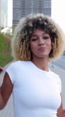 City Street 'te dans eden mutlu Afro-Amerikan kadının dikey HD videosu. Binlerce yıllık etnik kızın kameraya doğru giderken telefonda dans müziğinin tadını çıkarması ön planda.