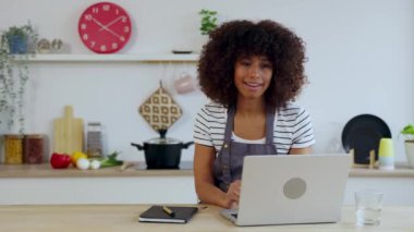 Genç Afro-Amerikalı güzel bir kadın evde çalışıyor ve yemek pişirirken dizüstü bilgisayarla konuşuyor. Seyahat ve iş konsepti