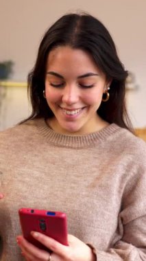 Modern ve şık bir mutfakta akıllı telefonunu karıştırıp rahat bir sabah rutininin tadını çıkaran genç bir kadının elinde bir fincan kahve tutarken çekilmiş dikey HD videosu. Yurtiçi yaşam tarzı ve sosyal medya kavramı