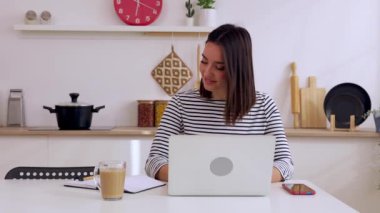 Genç, yetişkin bir kadın mutfakta dizüstü bilgisayar kullanırken not alıyor. Milenyum kadınlarının çevrimiçi sanal video dinlemeyi öğrenmesi. İş ve eğitim kavramı.