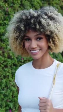 Şehir caddesinde yürürken kameraya gülümseyen genç ve çok ırklı bir kadının dikey HD videosu. Neşeli milenyum dişisi yazın açık havada yürüyüş yapıyor. İnsanlar ve mutluluk kavramı.