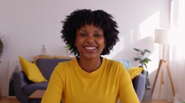 Dizüstü bilgisayar araması sırasında oturma odasında kameraya konuşan neşeli, genç Afrikalı Amerikalı bir kadın. Gülümseyen kadın arkadaşlarıyla ya da meslektaşlarıyla sanal online görüşme yoluyla sohbet ediyor..