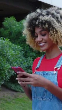 Latin kökenli genç bir kadının şehir caddesinde cep telefonu kullanırken çekilmiş dikey HD videosu. Z Gen kızı yazın dışarıda akıllı telefon uygulamasından mesaj yolluyor. Teknoloji yaşam tarzı ve sosyal medya kavramı.