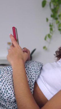 Dikey HD video. Sabahları yatakta dinlenirken cep telefonu kullanan tanınmayan genç bir kadının yan görüntüsü. Teknoloji ve ev hayatı