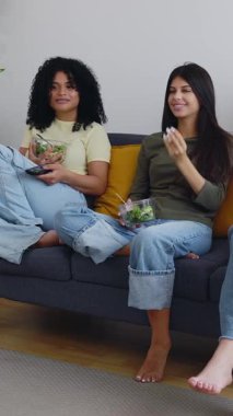 Evdeki kanepede oturmuş sağlıklı yemekler yerken televizyon izleyen üç güzel kadının dikey HD videosu. Boş zaman, arkadaşlık ve ev hayatı konsepti.