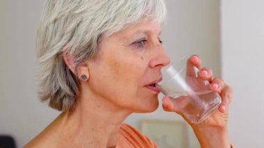 Sabah uyandıktan sonra evde kadehten su içen yaşlı bir kadın var. Sağlıklı yaşam tarzı kavramı.