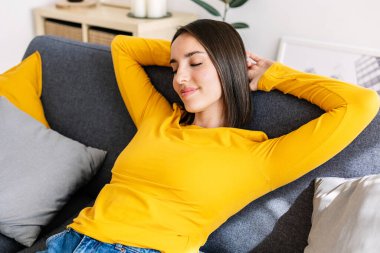 Genç, güzel bir kadın. Elleri başının arkasında. Parlak, modern bir dairede rahat bir kanepede dinleniyor. Sakinlik ve refah kavramı.