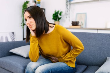 Karın ve diş ağrısı çeken, karnını ve yüzünü acılı bir ifadeyle tutan, oturma odasındaki bir kanepede oturan bir kadın.