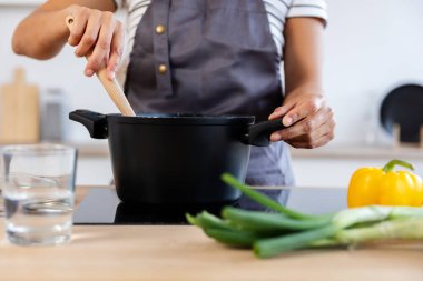 Kadın aşçı indüksiyon sobasında çorba pişirirken tahta kaşık kullanıyor, modern mutfağın yanında taze sebzeler var.