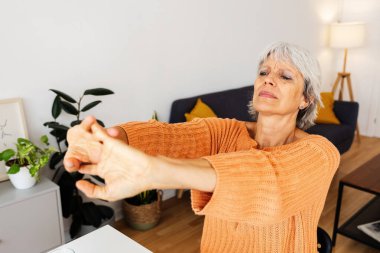 Üst düzey bir kadın rahat ofis masasında kollarını uzatıyor, evden işe gidip geliyor, rahatlıyor ve rahat bir oturma odasında bitkiler ve doğal ışıkla sağlıklı bir yaşam sürüyor.