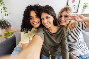 Üç bayan arkadaş evlerindeki kanepede otururken selfie çekiyorlar, gülümsüyorlar ve barış işareti yapıyorlar. Kadın arkadaşlığı ve gençlik kavramı