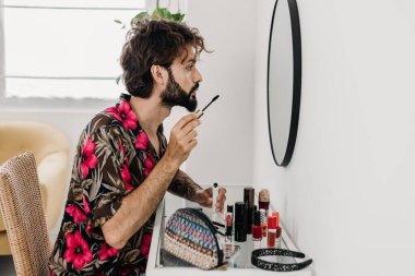 Renkli bir gömlek giyen genç sakallı bir adam makyaj masasında oturup aynaya bakarken maskara sürüyor. LGBTQ yaşam tarzı kavramı