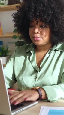 Profesyonel Latin Amerikalı iş kadınının parlak ofis içinde dizüstü bilgisayarda çalışırken çekilmiş dikey HD videosu. Gülümseyen siyah kadın işçi şirket grafik büyümesini analiz ediyor.